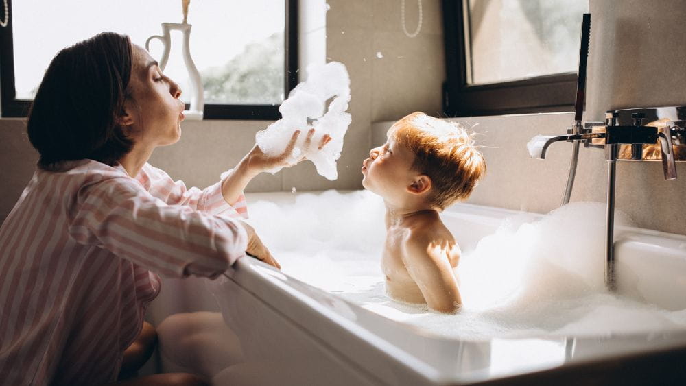 Young boy in bath blowing bubbles with mother.