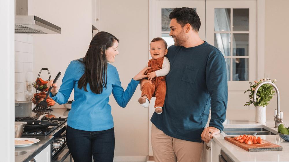 Dad holding child and mum cooking dinner in kitchen