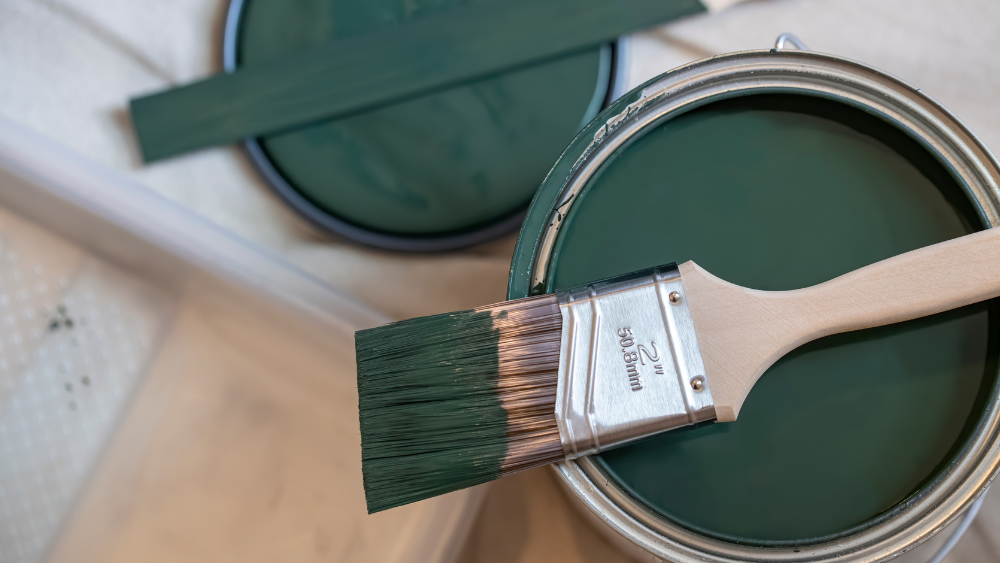 Open paint can with dark green paint and a paint brush laying across the top.